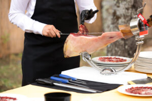 The chef is cutting the ham. image of a ham cutter with a knife cutting a thin slice of ham. Dried Ham, Serrano Ham, Acorn, Iberian, Italian or Parma Raw Prosciutto